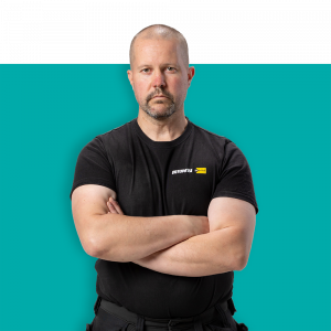 Vuokraamopäällikkö seisoo kädet ristissä, katsoen suoraan kameraan sinisellä taustalla.