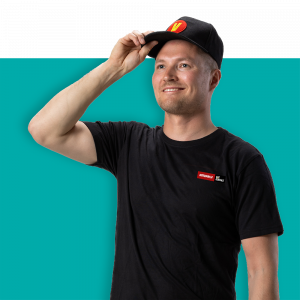 Mies mustassa t-paidassa ja lippiksessä hymyilee, pitäen lippiksen reunasta kiinni.