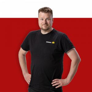 Mies mustassa t-paidassa, jossa on logot, seisoo punaisella taustalla kädet lanteilla.