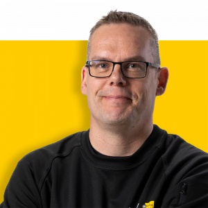 Läheltä katsottu kuva miehestä, jolla on päällä musta paita ja silmälasit, hymyilemässä keltaista taustaa vasten.